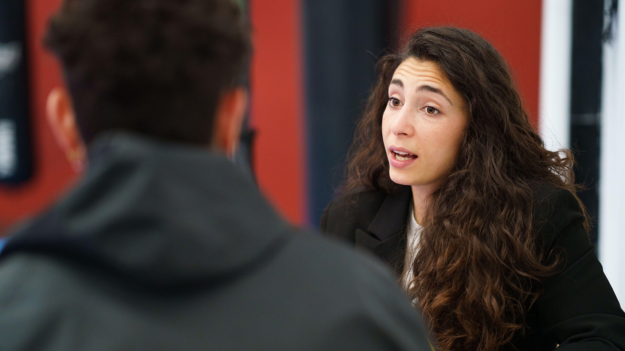 A curly haired woman speaking to another person who we see only the back of the head